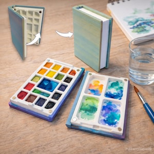 May include: A watercolor paint palette in a book-shaped case. The open palette displays a variety of vibrant watercolor paints. The case is blue and white, with a magnetic closure. A paintbrush and a glass of water are also visible.