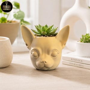 May include: A tan-colored Chihuahua-shaped planter holding a green succulent. The planter has a detailed face and large ears. The image also shows other small plants and the text "Pukka Printables".