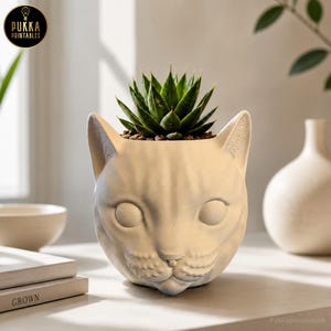 May include: A cream-colored cat-shaped planter with a succulent plant. The planter has a detailed cat face design with visible ears, eyes, and a nose. The succulent has green, pointed leaves. The text "Pukka Printables" is visible.