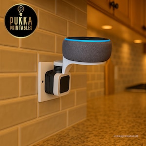 Pode incluir: Uma coluna inteligente cinzenta com um anel de luz azul está montada num suporte de tomada de parede branco. O suporte está ligado a uma tomada de parede. A coluna está posicionada acima de uma bancada de cozinha. O logótipo Pukka Printables está no canto superior esquerdo.