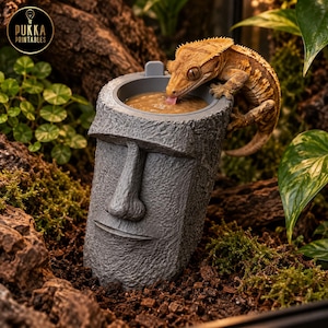 Puede incluir: Un cuenco de comida gris con textura de piedra, con forma de cabeza de estatua Moai y un recipiente circular en la parte superior. Un pequeño gecko crestado de color naranja amarronado está posado en el borde lamiendo la pasta marrón.