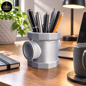 May include: A silver-colored, industrial-style pipe fitting pen holder with a threaded side opening. The cylindrical container holds several pens and a yellow pencil. The surface has a metallic finish with visible horizontal texture lines.
