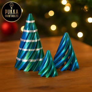 May include: Three decorative Christmas trees in shades of blue and green, with a spiral design. The largest tree has silver stripes. The trees are arranged on a wooden surface. The logo "Pukka Printables" is in the upper left corner.