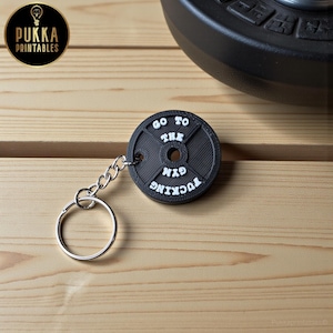 May include: A black weight plate keychain with the text "GO TO THE FUCKING GYM" in white. The keychain has a silver chain and ring. The background is a wooden surface with a black weight plate.
