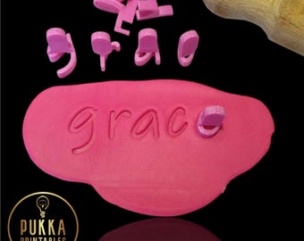 May include: Pink plastic letter stamps spelling the word "grace" on a pink play dough surface. The stamps are scattered around the play dough. A wooden rolling pin is in the background.