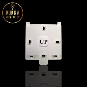 以下が含まれることがあります： 黒い文字で「UP」と印刷された白いプラスチック部品。この部品には、いくつかの楕円形と長方形の穴があり、黒い表面に反射しています。「Pukka Printables」のロゴが左上にあります。