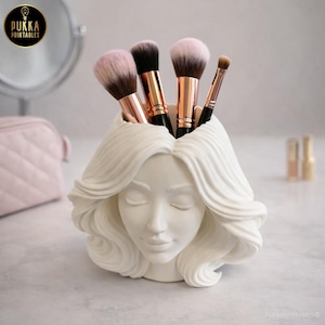 Peut inclure: Un porte-pinceaux de maquillage blanc en forme de tête sculptée, avec les yeux fermés et des cheveux ondulés. Le support contient plusieurs pinceaux de maquillage avec des poils roses et noirs et des manches en or rose. Le logo "Pukka Printables" est visible.