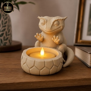 May include: A beige, textured figurine of a crested gecko with closed eyes and raised front paws, positioned behind a circular, geometric-patterned candle holder containing a small, glowing, warm-toned LED tea light candle.
