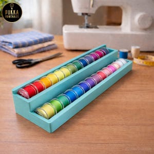 Op de afbeelding: Twee lichtblauwe houten garen organizers gevuld met kleurrijke spoelen garen. De spoelen zijn gerangschikt in een regenboogpatroon. Een naaimachine, schaar en stof staan op de achtergrond.