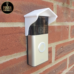 Op de afbeelding: Een Ring video deurbel bevestigd aan een rode bakstenen muur, beschermd door een witte plastic hoes. De deurbel is zilverkleurig met een zwarte cameralens en een wit verlichte knop. Het logo "Ring" is zichtbaar.