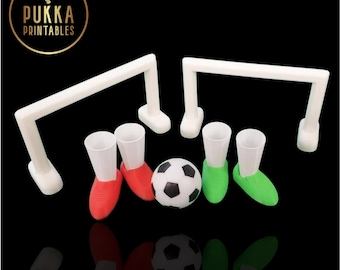 Op de afbeelding: Een voetbal-thema spelset met twee witte doelpalen, twee paar rood-groene schoenvormige shotglaasjes en een zwart-witte voetbal. Het logo "Pukka Printables" staat in een gouden cirkel.