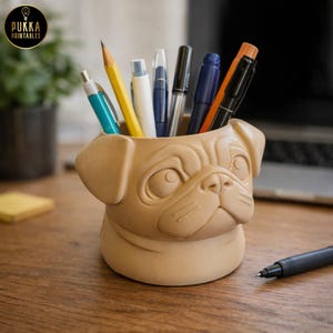 May include: A tan-colored desk organizer shaped like a pug dog's face, filled with pens and pencils. The organizer has detailed facial features and is sitting on a wooden desk. The logo "Pukka Printables" is in the upper left corner.