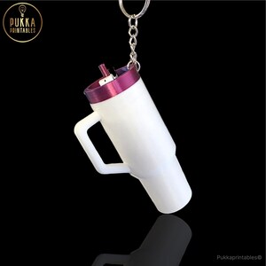 Op de afbeelding: Witte tumbler sleutelhanger met een paarse rand en rietje, bevestigd aan een zilveren sleutelhanger. De tumbler heeft een handvat. De tekst "Pukka Printables" is zichtbaar.