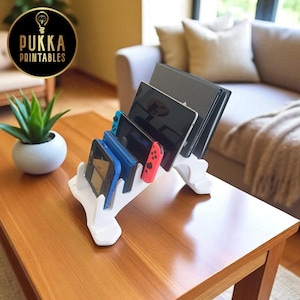 May include: A white, multi-device stand holding various electronics, including a tablet, Nintendo Switch, and other handheld gaming consoles. The stand is on a wooden table, with a plant and a couch in the background. The stand is made by Pukka Printables.