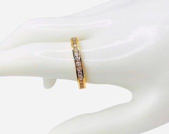 May include: A gold-colored ring with a band of small, clear gemstones. The ring is set on a white surface, showcasing its design. The gemstones are evenly spaced around the ring's circumference, creating a sparkling effect.