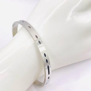 May include: A silver-toned metal cuff bracelet with a series of small, evenly spaced, dark oval cutouts. The bracelet is worn on a white mannequin hand, showcasing its simple, modern design. This minimalist jewelry piece is suitable for everyday wear.