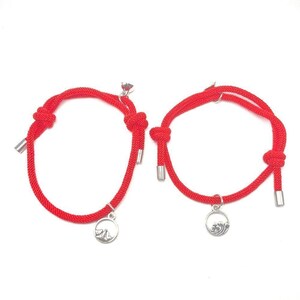 Puede incluir: Dos pulseras de cuerda roja con detalles y dijes plateados. Cada pulsera tiene un nudo y un tubo de metal en los extremos. Un dije presenta un diseño de montaña, y el otro un diseño de ola. Las pulseras son probablemente para parejas o amigos.
