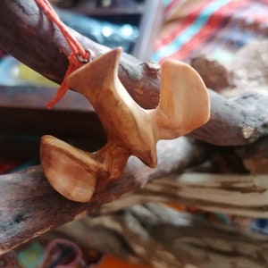 May include: A wooden pendant shaped like a bird in flight, with a natural wood finish. The pendant is hanging from a red cord.