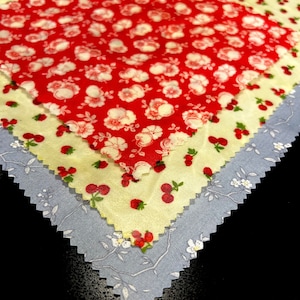 Peut inclure: Gros plan de carrés de tissu superposés avec divers motifs floraux et fruités. La couche supérieure est rouge avec des fleurs blanches, suivie de jaune avec des cerises rouges, et d'une couche bleu clair avec des fleurs blanches.