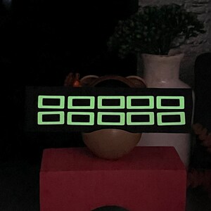 Puede incluir: Una diadema negra con ocho cuadrados verdes brillantes. Los cuadrados están dispuestos en dos filas de cuatro.