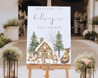 May include: A white rectangular welcome sign on a wooden easel featuring the text Welcome to Kelsey's Baby Shower. The illustration depicts a snow-covered log cabin, green pine trees, a deer, a raccoon, a hedgehog, and a bear.