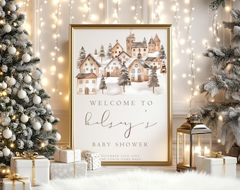 May include: A gold-framed sign featuring a watercolor illustration of a snowy village with houses and trees. The text reads: WELCOME TO kelsey's BABY SHOWER DECEMBER 24TH 20XX THE VENUE NAME HERE. The sign is displayed in a festive setting.