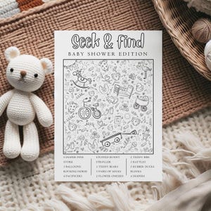 Peut inclure: Un jeu "Seek & Find Baby Shower Edition" en noir et blanc avec des illustrations d'articles pour bébés. Un ours en peluche blanc au crochet est à côté du jeu. Le jeu comprend une liste d'articles à trouver, tels que des couches, des hochets et des canards en caoutchouc.