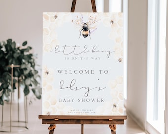 May include: A wooden easel displays a baby shower welcome sign. The sign features the text "little honey is on the way" and "Welcome to Kelsey's Baby Shower." The design includes a bee illustration and honeycomb pattern. The sign is propped up on a wooden easel.