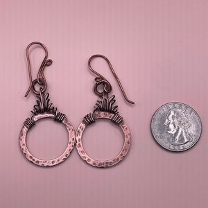 Op de afbeelding: Handgemaakte koperen oorbellen met een gehamerde textuur en decoratieve draad details. De oorbellen hebben een cirkelvormig ontwerp met een bloemmotief aan de bovenkant. Een munt ligt naast de oorbellen ter vergelijking van de grootte.