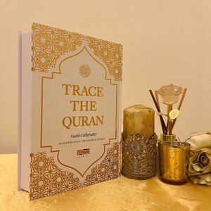 May include: A white book with gold lettering titled "Trace the Quran" with the subtitle "Naskh Calligraphy Handwritten Traceable Quran". The book is on a gold table with a gold candle and a gold diffuser with a glass rose.