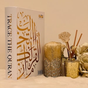 May include: A white book with gold lettering that says "TRACE THE QURAN" on the spine. The front cover features gold Arabic calligraphy. The book is next to a gold candle holder with a gold candle and a gold diffuser with a clear glass bottle and gold reeds.