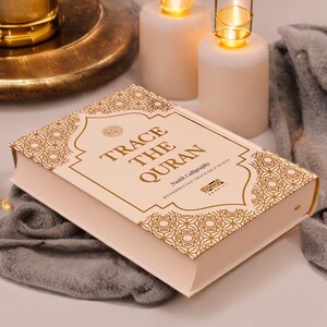May include: A pink book with gold lettering that says "Trace the Quran" and "Naskh Calligraphy" with a gold patterned design on the cover.