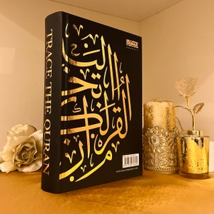 May include: A black hardcover book with gold lettering on the cover. The book is titled "Trace the Quran" and features Arabic calligraphy in gold. The book is on a gold-colored surface.