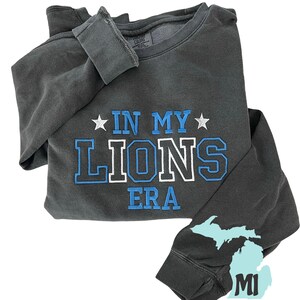 May include: A charcoal gray sweatshirt with the text "In My Lions Era" in blue and white lettering. The sweatshirt has a Michigan state outline on the sleeve with the letters "MI" and the text "Custom Gifts".