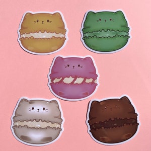 May include: Five cat-shaped macaron stickers. The macarons are in different colors: yellow, green, purple, beige, and brown. Each macaron has a cute cat face with black eyes and whiskers.