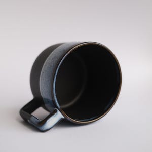 Pode incluir: Uma caneca de cerâmica azul escura com um esmalte azul claro na alça e na borda. A caneca está vazia e está sobre uma superfície branca.