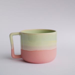 Może przedstawiać: Ceramiczny kubek w kolorze jasnozielonym i różowym z zaokrąglonym uchwytem. Kubek ma jasnozielony wierzch i różowe dno.
