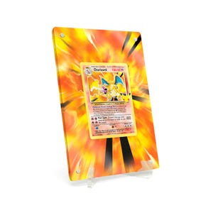 Può includere: Una carta Pokémon Charizard incorniciata. La carta è centrata su uno sfondo di fiamme arancioni e gialle. La carta presenta una creatura simile a un drago giallo e arancione con ali blu. La cornice è trasparente e sostenuta da un supporto trasparente.