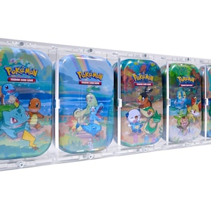 Op de afbeelding: Vijf Pokémon Trading Card Game-blikken in een transparante acryl display. Elke blik heeft een ander Pokémon-personage op een blauwe en groene achtergrond. De blikken zijn gelabeld "Pokémon Trading Card Game".