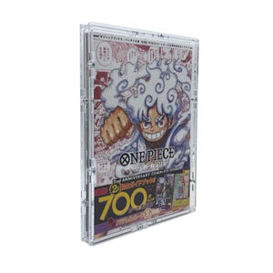 Può includere: Una vetrina trasparente in acrilico che contiene una guida al gioco di carte One Piece. La copertina presenta un'illustrazione vibrante di un personaggio con i capelli bianchi e un'espressione determinata. Il testo include "ONE PIECE CARD GAME" e "2nd ANNIVERSARY COMPLETE GUIDE".