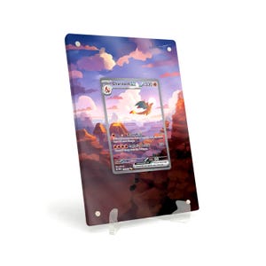 Puede incluir: Una tarjeta Pokémon Charizard EX enmarcada, exhibida en un soporte acrílico transparente. La tarjeta presenta una ilustración de un dragón naranja llameante contra un paisaje volcánico. El arte de fondo representa una puesta de sol con tonos rosas y morados.