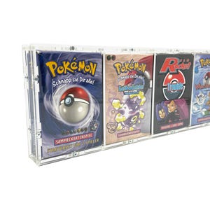 May include: A clear acrylic display case containing four rectangular Pokémon trading card game boxes. The boxes feature colorful graphics, including Poké Ball imagery and various character illustrations, with text such as Pokémon, Team Rocket, and Sammelkartenspiel.