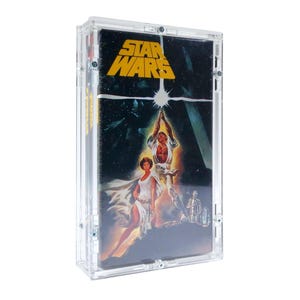 Peut inclure: Un boîtier en acrylique transparent contenant une cassette VHS de Star Wars. Le boîtier a un fond noir avec un titre "Star Wars" jaune et une scène du film mettant en scène Luke Skywalker, la princesse Leia et Dark Vador.