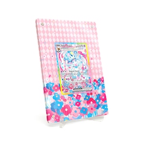 Peut inclure: Un présentoir rose et blanc avec une carte Pokémon Sylveon Gx. Le présentoir présente un motif de losanges roses et un motif floral en bas. La carte présente une illustration colorée de Sylveon avec le texte "Magical Charm".