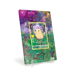 Puede incluir: Una carta coleccionable de Pokémon Pikachu, exhibida en un soporte acrílico transparente. La carta presenta un personaje Pikachu amarillo con el texto "Pikachu". El fondo es una colorida escena de bosque con tonos morados y verdes.