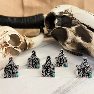 May include: Five silver rings with turquoise stones and engraved letters A, B, C, D, and E. The rings are shaped like cattle ear tags.