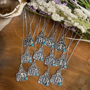 May include: A collection of silver necklaces with turquoise stones. Each necklace features a silver pendant shaped like a cattle ear tag with a letter engraved on it.