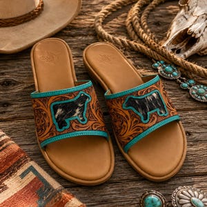 May include: A pair of tan leather slide sandals featuring a tooled floral pattern, turquoise trim, and a black and white cow-shaped hair-on-hide inlay on the wide strap. The footbed is smooth tan leather with a subtle embossed logo detail.