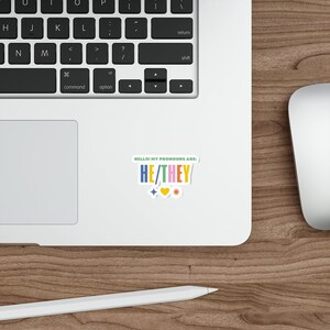 Peut inclure: Un autocollant avec le texte "HELLO! MY PRONOUNS ARE: HE/THEY" en lettres colorées sur un fond vert. L'autocollant comprend également un cœur rose, une étoile jaune et un diamant bleu.