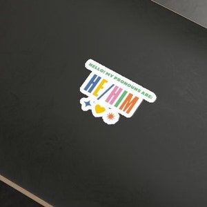 May include: A white sticker with colorful text that reads "Hello! My pronouns are: He/Him". The sticker has a heart and two stars.